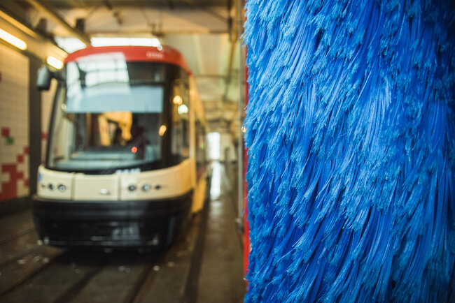 Na pierwszym planie widać zbliżenie na pionową, niebieską szczotkę obrotową, która jest częścią automatycznej myjni dla pojazdów wielkogabarytowych. W tle, w głębi hali, znajduje się nowoczesny, niskopodłogowy tramwaj oczekujący na czyszczenie.