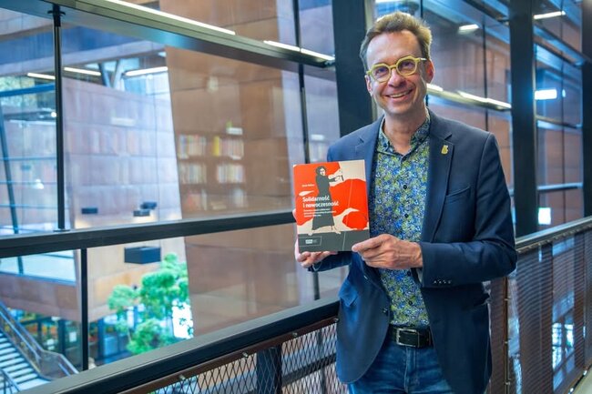 Szczupły mężczyzna w okularach pozuje z książką do zdjęcia 