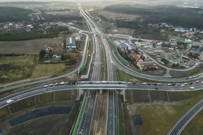 Na zdjęciu widać z lotu ptaka rozbudowany węzeł drogowy z nowoczesną drogą ekspresową oraz przebiegającym nad nią wiaduktem. Wokół trasy znajdują się tereny zabudowane, pola oraz place budowy, co wskazuje na niedawno zakończoną lub trwającą inwestycję infrastrukturalną. 