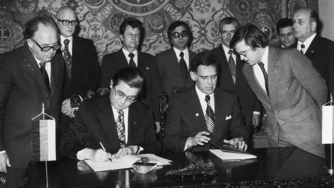 Czarno-białe zdjęcie przedstawia grupę mężczyzn w garniturach podczas oficjalnej uroczystości podpisywania dokumentów przy dużym stole. W centrum uwagi znajdują się dwie siedzące postacie skupione na formalnościach, którym z powagą przyglądają się pozostali uczestnicy wydarzenia.