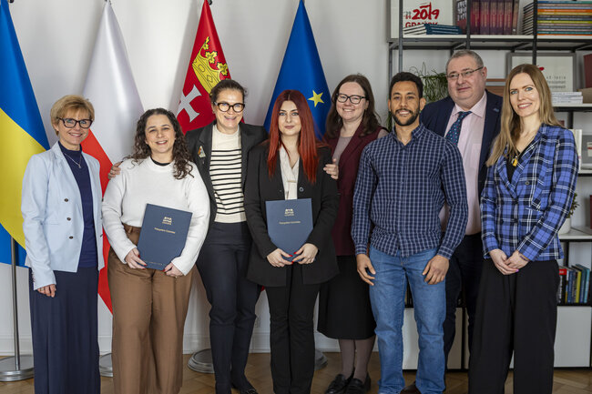 Sześć kobiet i dwóch mężczyzn pozuje do zdjęcia w gabinecie na tle flag. Uśmiechają się.