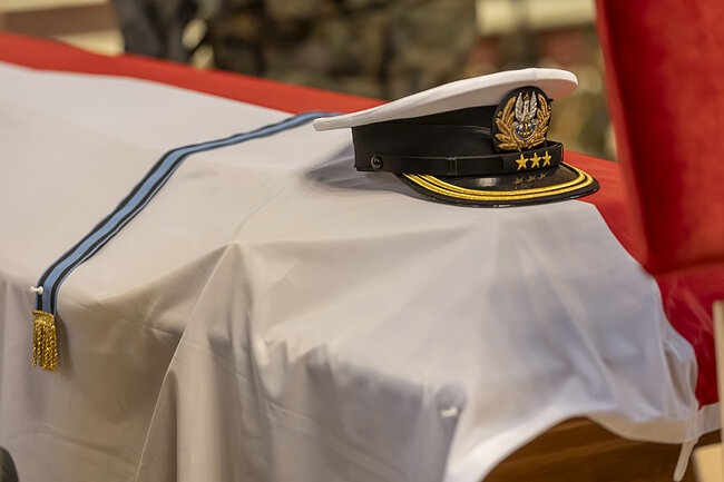 Zdjęcie przedstawia czapkę oficera Marynarki Wojennej ułożoną na trumnie przykrytej biało-czerwoną flagą. Obok nakrycia głowy widoczna jest błękitna wstęga orderowa, co wskazuje na uroczysty charakter ceremonii pogrzebowej z honorami wojskowymi. 