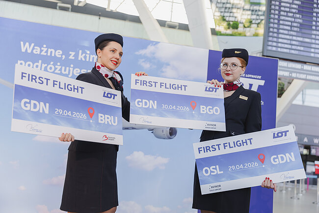 Zdjęcie przedstawia dwie uśmiechnięte stewardessy linii lotniczych LOT trzymające pamiątkowe tablice upamiętniające pierwsze loty między Gdańskiem a Brukselą, Oslo i Bergen. Wydarzenie odbywa się w terminalu lotniska, co podkreślają tablice odlotów oraz eleganckie mundury personelu pokładowego.