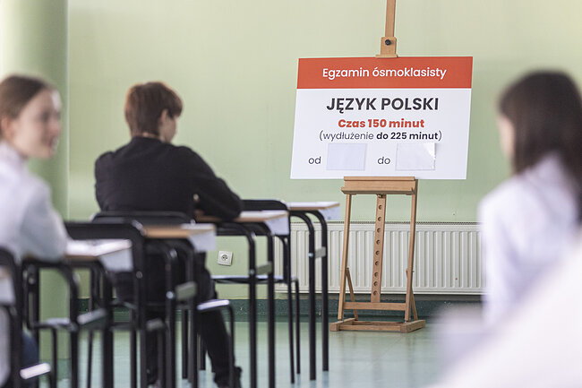 Uczniowie siedzą przy stolikach egzaminacyjnych - w tle, przy ścianie znajduje się tablica, z informacją że mają do dyspozycji 150 minut