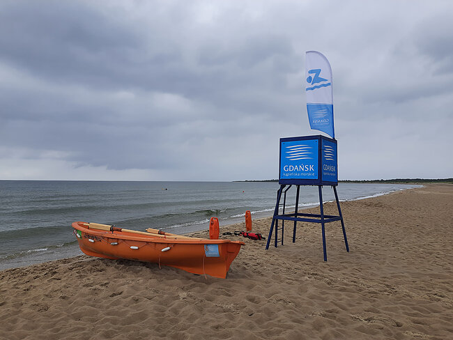 Fotografia dekoracyjna przedstawiająca plażę oznaczoną znakiem Gdańsk kąpieliska morskie oraz łódź ratowniczą