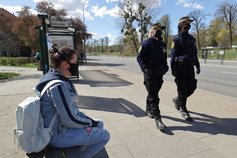 Na zdjęciu widać dwie strażniczki miejskie ubrane w służbowe mundury, z maseczkami ochronnymi na twarzach. Po lewej stronie zdjęcia na ławce siedzi młoda dziewczyna, także w maseczce ochronnej. Ma na sobie jasną bluzę dresową, dżinsy i jasny plecak. W tle widać wiatę przystanku autobusowego