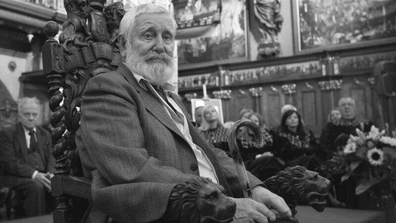 Starszy siwy mężczyzna z brodą elegancko ubrany siedzi w rzeźbionym fotelu 