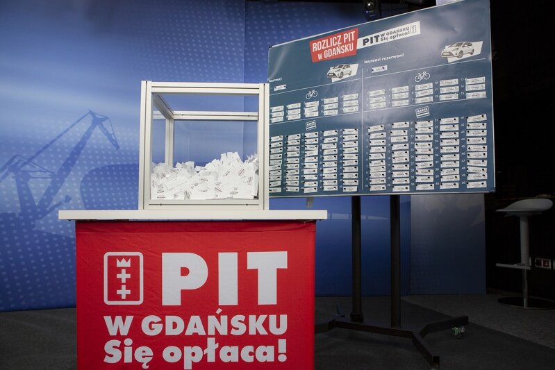 Szklany kwadrat w którym umieszczono losy, stoi na stoliku z napisem Pit w Gdańsku. Się opłaca W tle tablica z przyczepionymi kuponami. 