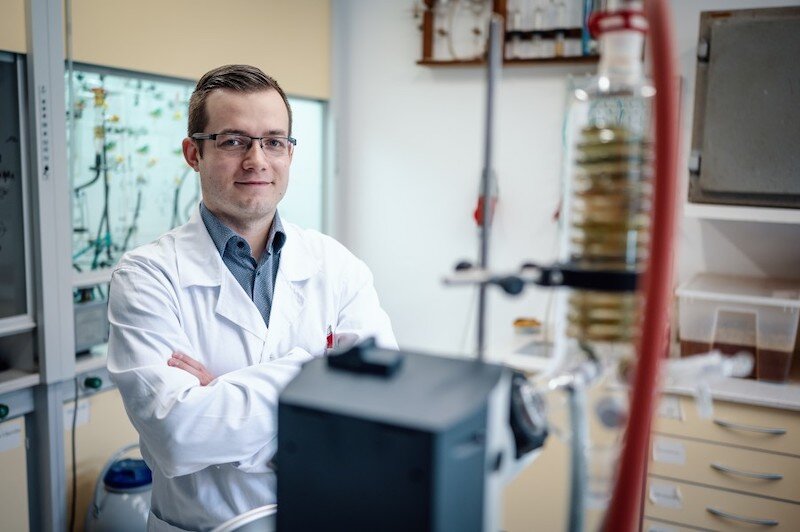 Mężczyzna w białym fartuchu i okularach stoi z założonymi rękoma i patrzy w obiektyw aparatu