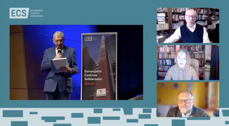 Czworo ludzi: troje mężczyzn i jedna kobieta. Zdjęcie przedstwia mężczyznę stojącego na scenie i troje rozmówców, którzy biorą udział w spotkaniu online, widoczne są ich głowy