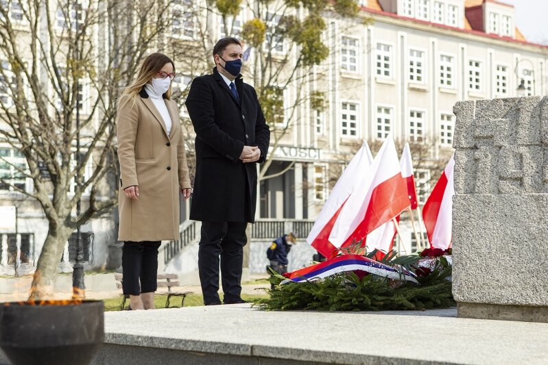 Kobieta i mężczyzna w maseczkach stoją przed pomnikiem. Przy pomniku wiązanka kwiatów. W tle budynki