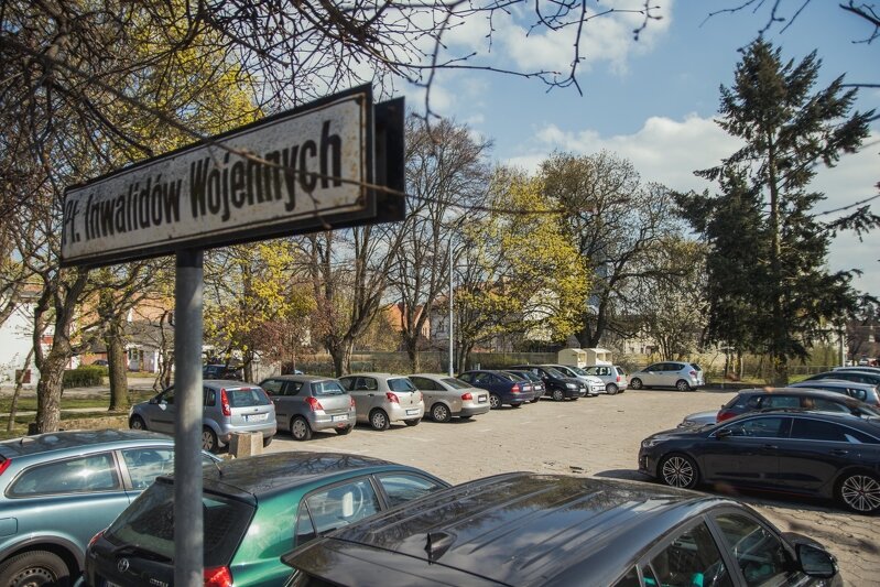 Plac na którym zaparkowanych jest kilkanaście samochodów. Z placem sąsiadują stare drzewa. Po lewej znajduje się metalowy słupek z poprzeczną białą tablicą, na której jest napis: Pl. Inwalidów Wojennych