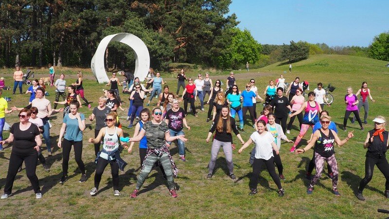 Kilkadziesiąt osób w strojach sportowych na świeżym powietrzu - stoją w rozkroku, mają ręce rozłożone na boki