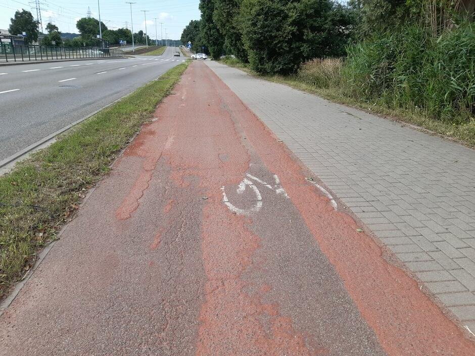 na zdjęciu fragment zniszczonej drogi rowerowej w kolorze czerwonym, widać wyraźne dziury, obok po lewej widać fragment jezdni