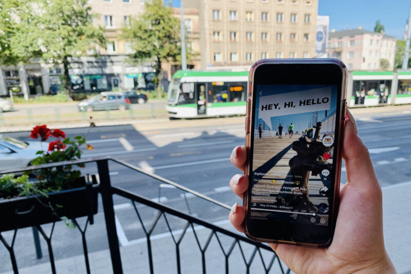 dłoń trzyma smartfona, na którym widać obrazek z napisem hey, hi, hello, w tle szeroka ulica i przejeżdżający tramwaj