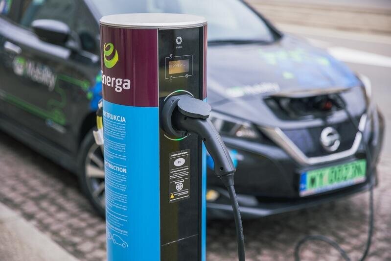 Zdjęcie przedstawia tzw. stację ładowania - jest to metalowy słupek, z którego wychodzi przewód zasilający akumulator samochodu elektrycznego. Podłączony samochód stoi za słupkiem