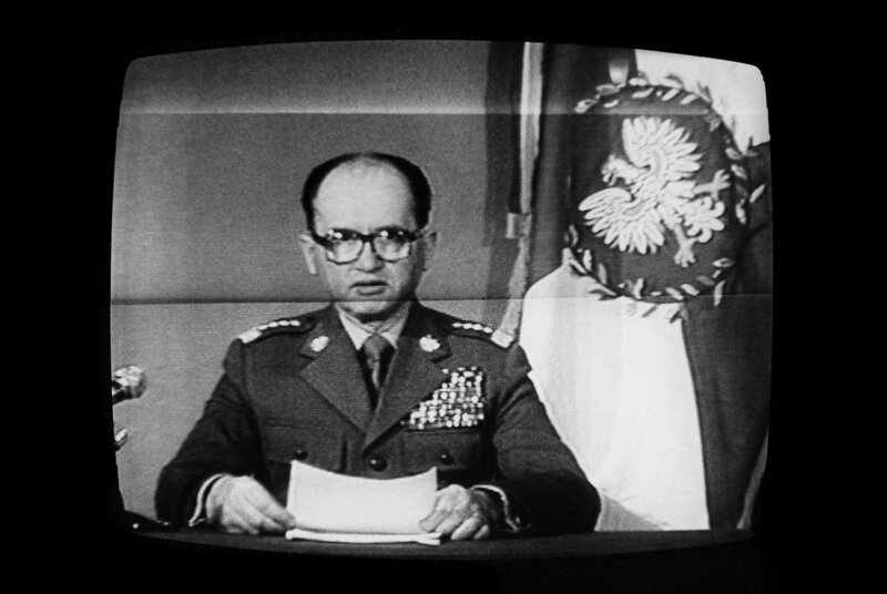 Czarno-białe zdjęcie: telewizor, na monitorze mężczyzna w wojskowym mundurze z kartka, w okularach, obok po lewej sztandar z godłem Polski