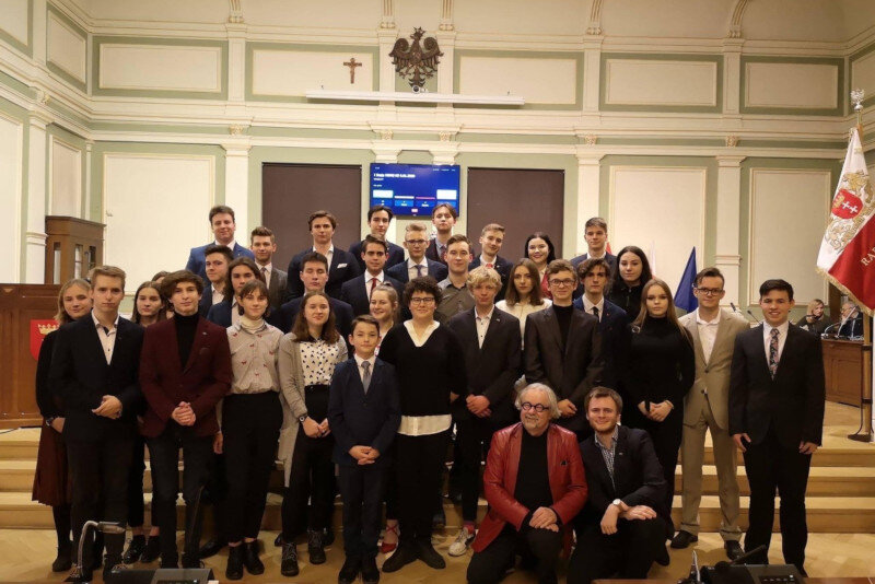 kilkadziesiąt osób młodych i starszych pozuje z uśmiechami na tle ściany z godłem i krzyżem