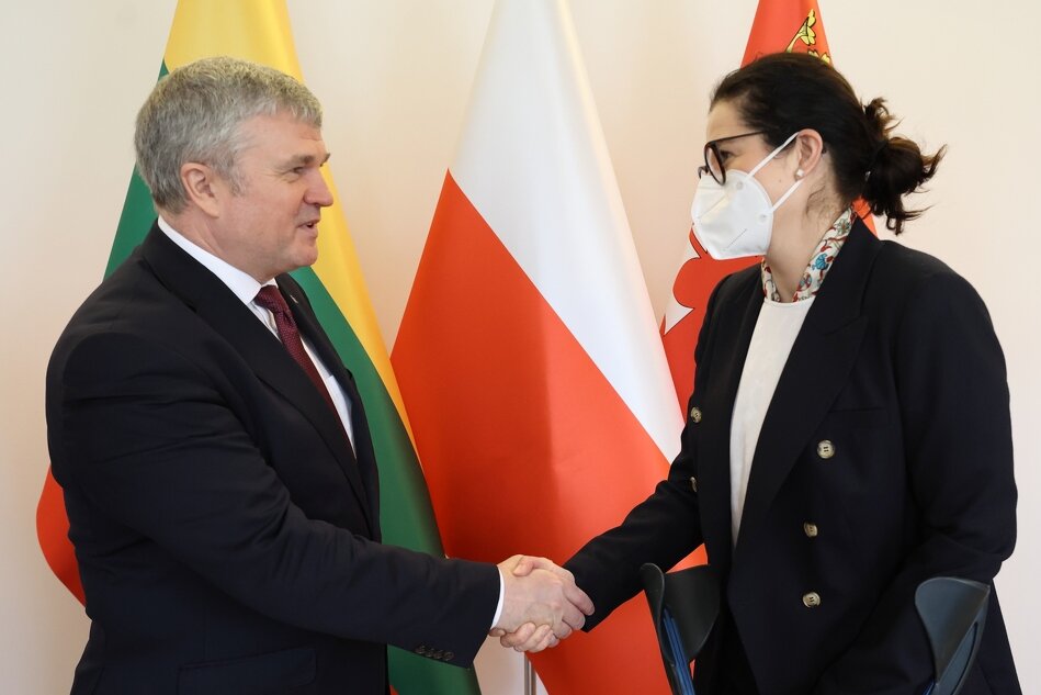 Ambasador Litwy i Prezydent Gdańska podają sobie rękę na powitanie podczas spotkania w Urzędzie Miejskim w Gdańsku. W tle są flagi, patrząc od lewej: Litwy, Polski i Gdańska. Ambasador Litwy jest bez maseczki sanitarnej - stoi po lewej stronie. Prezydent Gdańska jest w maseczce, stoi po prawej