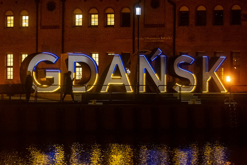 Napis GDAŃSK podświetlony na niebiesko (u góry) i żółto (na dole). Przed napisem rzeka z odbiciem napisu, w tle budynek