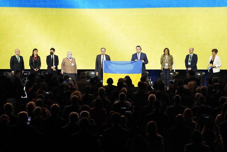 minuta cisz, sala obrad pełna ludzi, na scenie w garniturach dwóch mężczyzn trzyma niebiesko-zieloną flagę