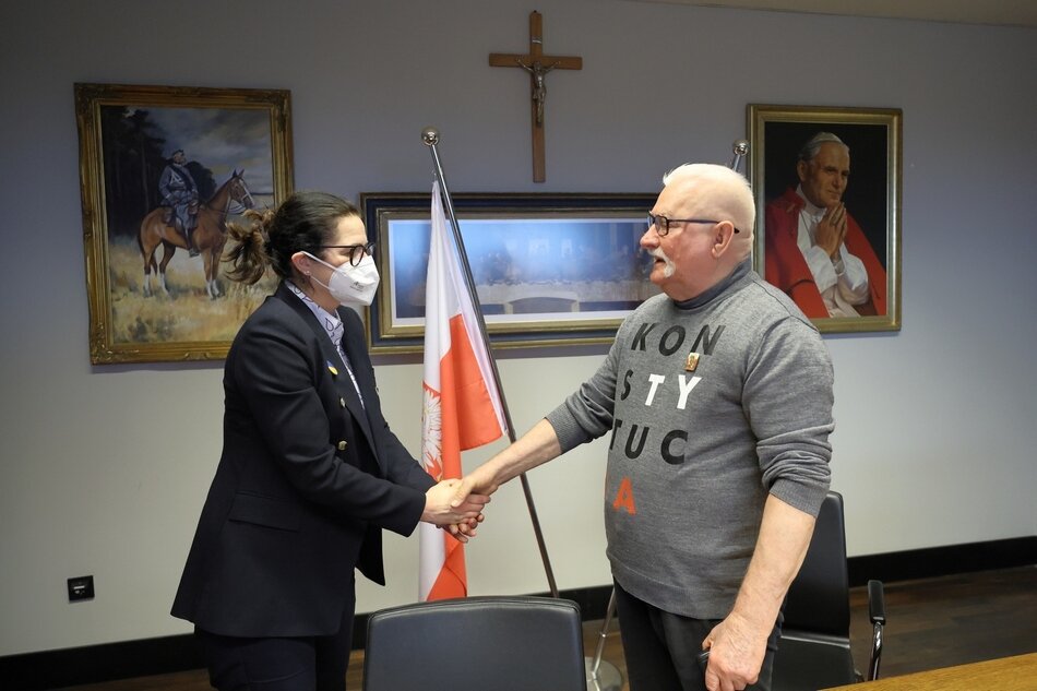 Kobieta w żakiecie, okularach i maseczce na twarzy wita się ze starszym mężczyzna w okularach z koszulce z długim rękawem z napisem KONSTYTUCJA. W tle ściana: na niej od lewej: obraz (Józef Piłsudski na koniu), krzyż, obraz z wizerunkiem św. papieża Jana Pawła II