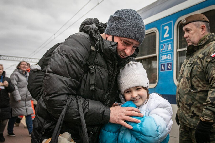 Peron na dworcu kolejowym, w tle niebieski wagon. Na pierwszym planie widzimy wzruszonego mężczyznę w kurtce i czapce, który pochyla się przytulając kilkuletnią dziewczynkę. Po prawej stoi mężczyzna w polskim zielonym mundurze i brązowym berecie 