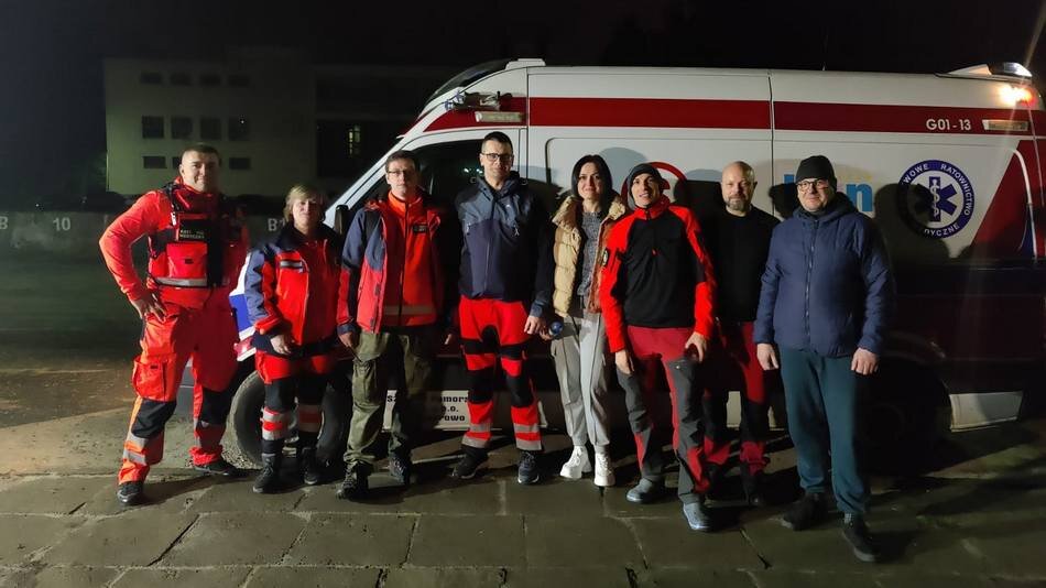 Ciemno. Osiem osób, większość w ubraniach ratowników medycznych stoi obok siebie. Za nimi karetka pogotowia