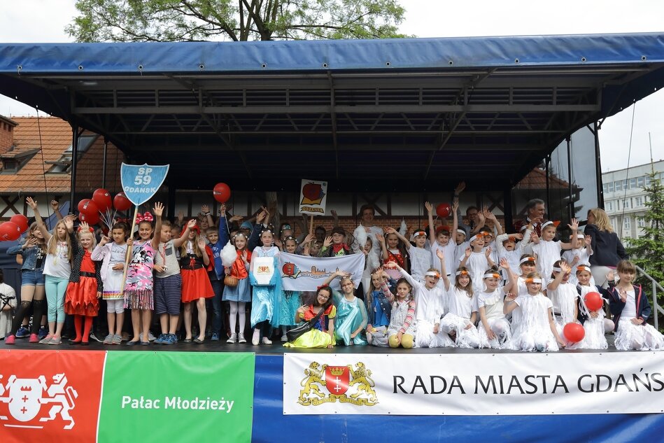 Duża grupa dzieci w wieku szkolnym stoi na plenerowej scenie. Pod nimi baner z napisem Rada Miasta Gdańska
