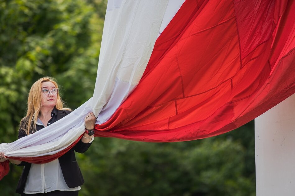Po lewej stronie jest kobieta, długowłosa blondynka w okularach, która trzyma w rękach wielką biało-czerwoną flagę. Ta flaga jest już przymocowana do linki potężnego masztu, który znajduje się z prawej strony. Za chwilę flaga zacznie swoją podróż w górę, wprost z rąk trzymającej ją kobiety 