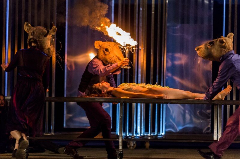 Scena teatralna, na środku łóżko, na nim leży naga śpiąca kobieta. Z prawej strony, w środku i z lewej trzech mężczyzn z pochodniami na główach mają maski szczurów 