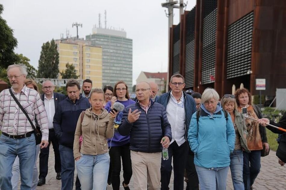 grupa osób idzie obok nowoczesnego budynku ECS, w tle trzy krzyże, w środku niewysoki starszy mężczyzna w okularach