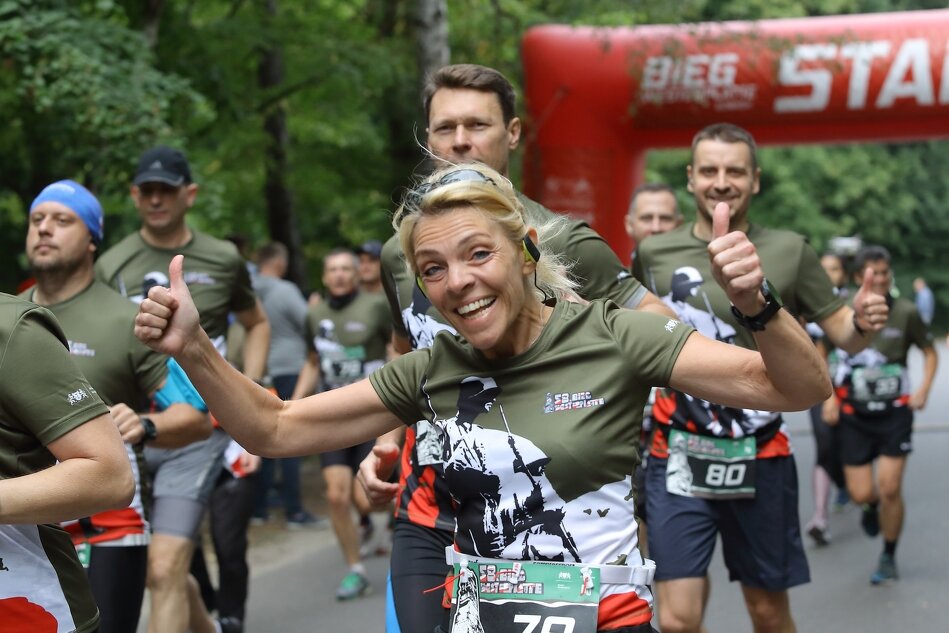 Biegnący ludzie w zielonych koszulkach. Na pierwszym planie uśmiechnięta blond kobieta z rozpostartymi rękoma. W tle napis START 