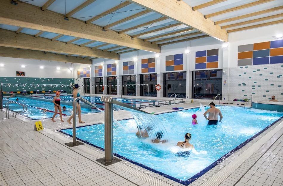 basen na którym e brodziku bawi się kilka osób dorosłych i dzieci, widać bicze wodne, w tle duża niecka i jacuzzi, w którym siedzi mężczyzna w czepku