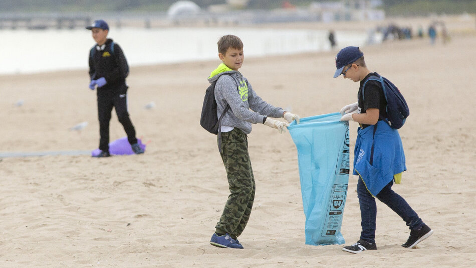 dzieci zbierają śmieci na plaży, na pierwszym planie dwóch chłopców trzyma duży niebieski worek na śmieci, za nimi trzeci chłopak niesie śmieci. 