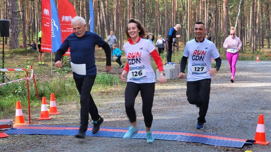 Starszy mężczyzna, młoda kobieta i młody mężczyzna wbiegają na metę. Wokoło zieleń