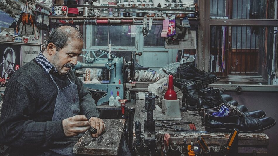 Mężczyzna w starszym wieku w zakładzie szewskim przy stole, obok lezą buty i różne narzędzia
