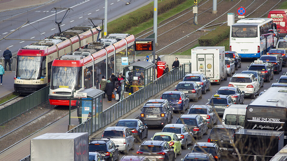 Z prawej strony sznur samochodów, jeden za drugim, w trzech rzędach. Z lewej dwa tramwaje na torach 