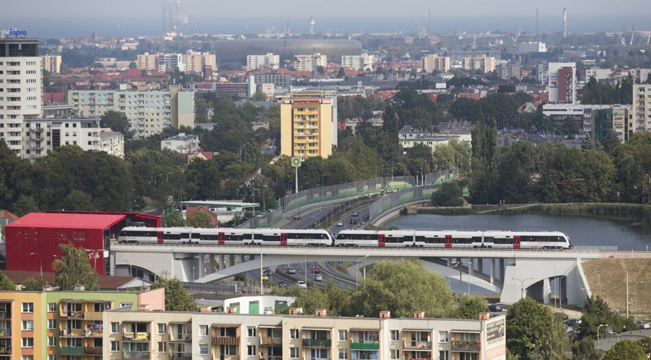 Na pierwszym planie na dole ostatnie piętro bloku mieszkalnego, wyżej wiadukt i jadący po nim pociąg, w tel widać bloki, wieżę kościoła, za nimi stadion sportowy