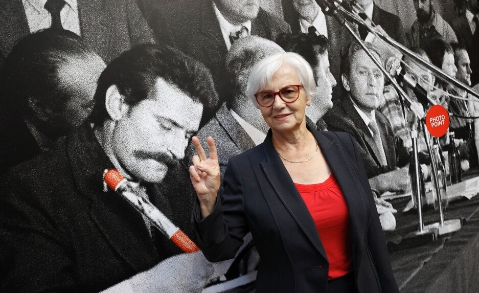 Kobieta w lekko przyciemnianych okularach pokazuje znak zwycięstwa na tle dużego zdjęcia z podpisania Porozumień Sierpniowych w Gdańsku w 1980 roku. Na pierwszym planie zdjęcia, po lewej, podpisujący Lech Wałęsa 