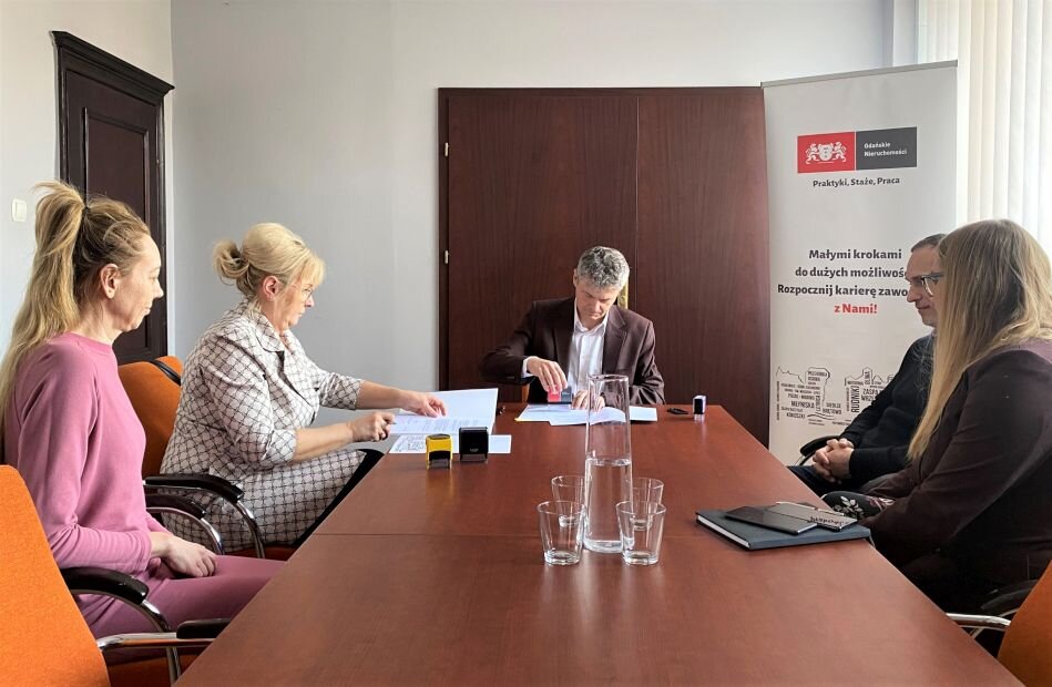 za stołem w pokoju biurowym siedzą 4 osoby, kobiety i mężczyźni i podpisują dokument