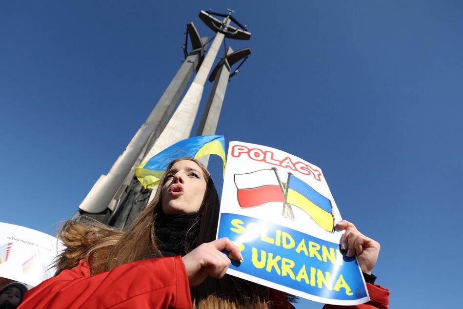 Młoda kobieta w czerwonej kurtce stoi pod Pomnikiem Poległych Stoczniowców, trzyma kartkę na której namalowane są flagi Polski i Ukrainy, a także napis: Polacy solidarni z Ukrainą. Za jej plecami, pod pomnikiem widać powiewającą flagę w barwach Ukrainy