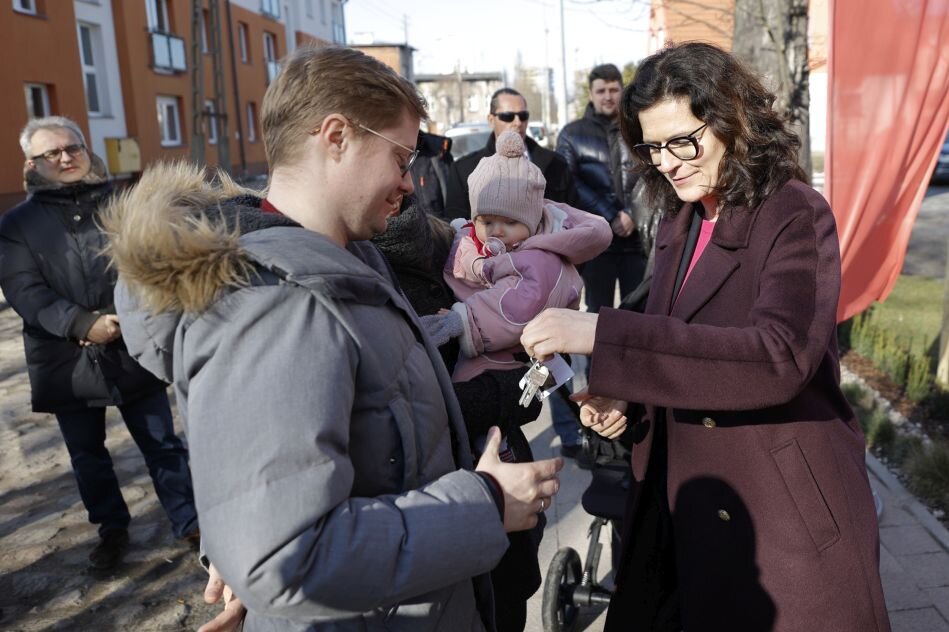 Kobieta w średnim wieku, w płaszczu trzyma klucze i przekazuje je rodzinie z dzieckiem na ręku
