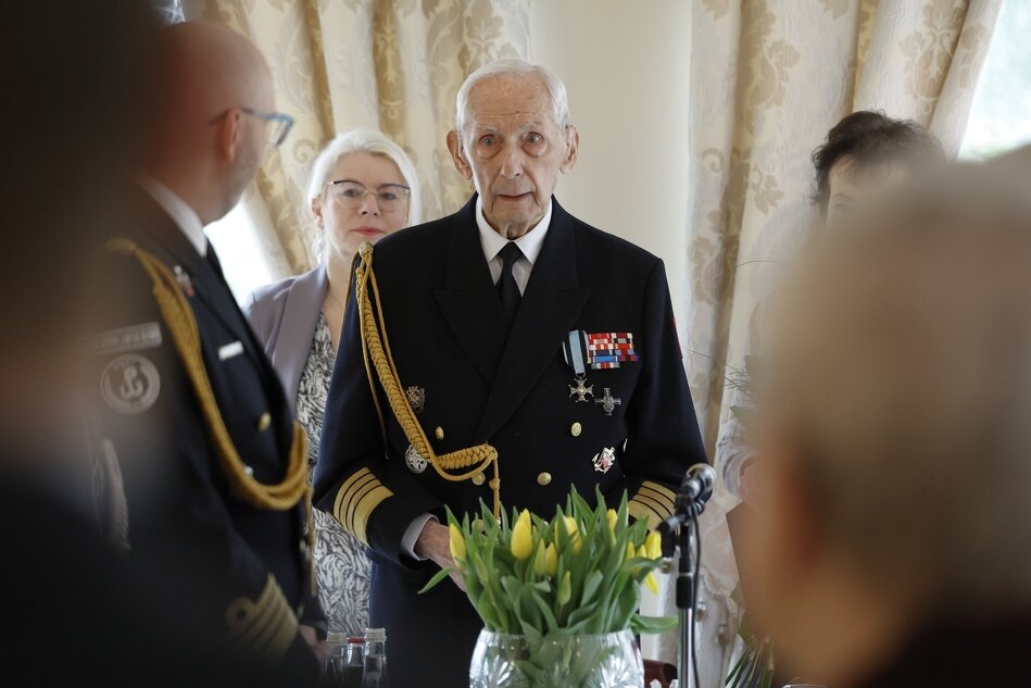 sędziwy mężczyzna z siwymi włosami, w mundurze z medalami stoi otoczony ludźmi przy stole