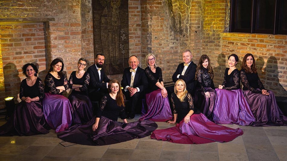12 osób (kobiety i mężczyźni) elegancko ubrani siedzą w starodawnym pomieszczeniu z cegieł