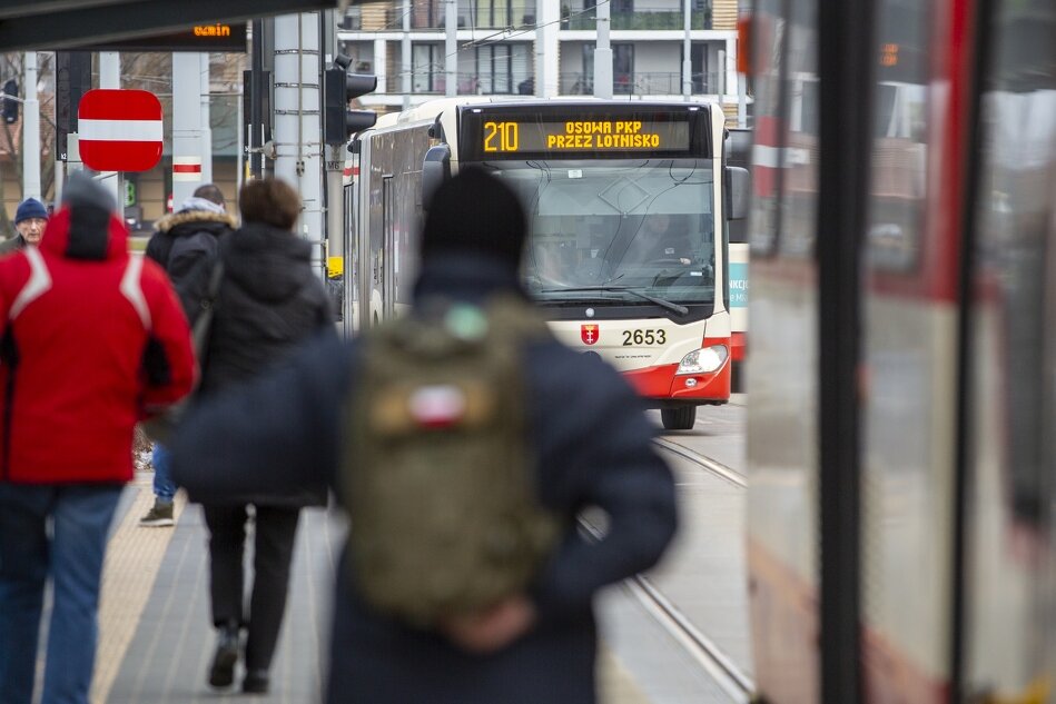 Zdjęcie przedstawia autobus miejski na przystanku i idące do niego osoby, których jest kilka