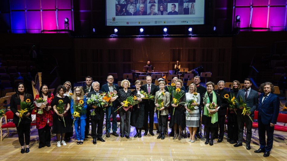 Grupa kilkudziesięciu osób elegancko ubranych, z kwiatami i statuetkami w dłoniach, stoi na scenie.