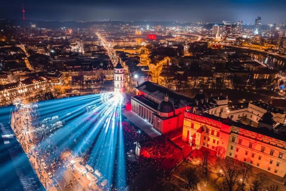 Nocna panorama miasta zrobiona z powietrza, prawdopodobnie dronem. Centralnie widzimy wieżę katedry wileńskiej, od której bija wielkie snopy niebieskiego światła. Po prawej widać również iluminacje w czerwonym kolorze