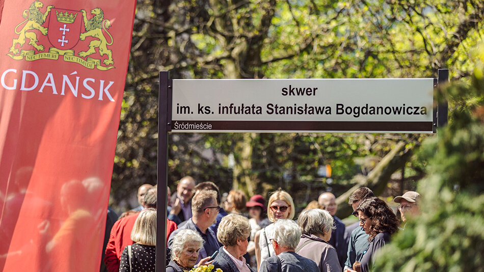 na pierwszym planie czerwona flaga z herbem Gdańska oraz tablica z nazwą skweru w tle grupa ludzi