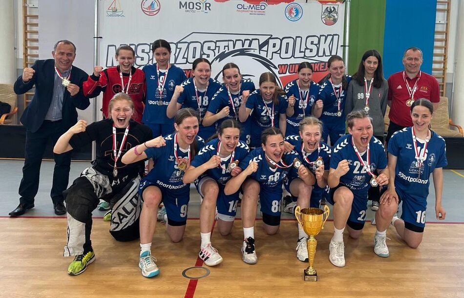 Zawodniczki w strojach sportowych w dwóch rzędach cieszą się, na szyjach mają medale. Po bokach stoją trenerzy 
